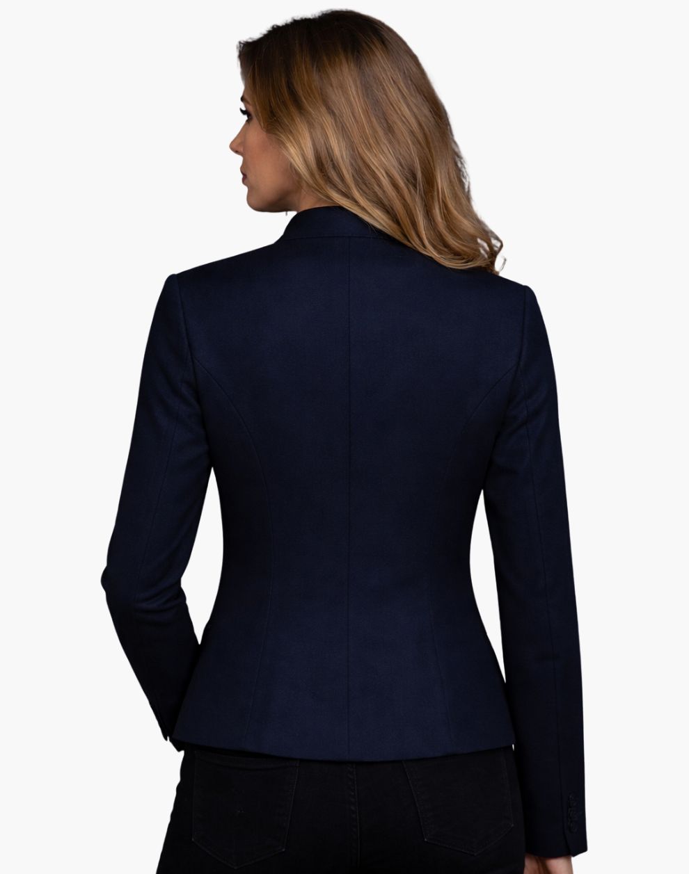 Back view of a woman wearing a fitted deep navy tailored jacket with clean vertical seams that contour the waist and a slightly flared hem. The structured jacket is paired with dark jeans, and her shoulder-length blonde hair falls over the collar. The background is plain light gray in a studio setting.