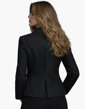 Back view of a woman wearing a fitted dark wool jacket with a structured silhouette and a center back vent. The jacket features a deep green collar accent and subtle shaping seams that contour the waist. She pairs it with dark jeans, and her shoulder-length blonde hair falls loosely over the collar. The background is plain light gray in a studio setting.