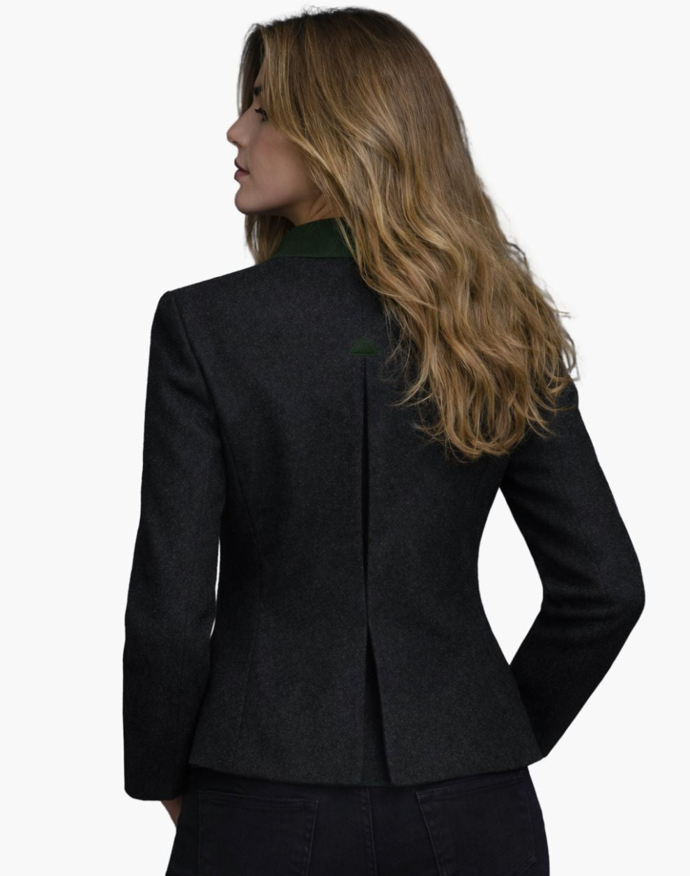 Back view of a woman wearing a fitted dark wool jacket with a structured silhouette and a center back vent. The jacket features a deep green collar accent and subtle shaping seams that contour the waist. She pairs it with dark jeans, and her shoulder-length blonde hair falls loosely over the collar. The background is plain light gray in a studio setting.