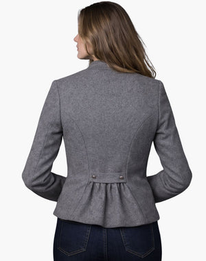 Back view of a woman wearing a light grey tailored Trachten jacket with a fitted waist and pleated back detail, paired with dark jeans, against a white background.