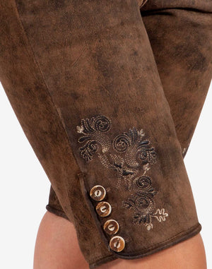 Close-up of brown leather women’s lederhosen leg showing traditional embroidered floral design and decorative side buttons on a textured vintage-style leather finish.