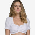 Front view of a woman wearing a white dirndl-style cropped blouse with short puff sleeves, lace trim neckline, front tie detail, and elastic hem.