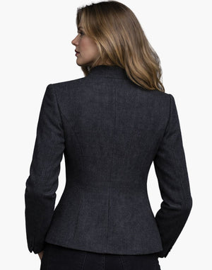 Back view of a woman wearing a fitted charcoal grey tailored jacket with long sleeves and structured seams that contour at the waist. The hem flares slightly over dark jeans, and her shoulder-length light brown hair falls naturally over the collar. The background is plain light gray, creating a clean studio setting.
