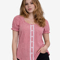Front view of a woman wearing a red and white gingham dirndl-style blouse with short puff sleeves, lace front panel, and decorative buttons, paired with black denim shorts.