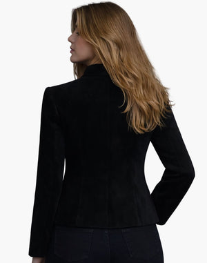 Back view of a woman wearing a fitted black tailored jacket with long sleeves and structured seams that shape the waist. The hem flares slightly over dark jeans, and her shoulder-length blonde hair falls loosely over the collar. The background is plain light gray in a studio setting.