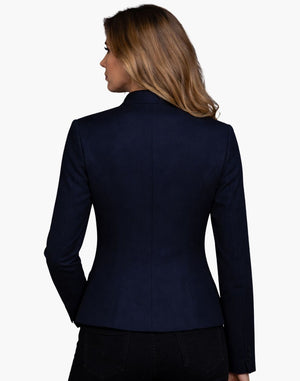Back view of a woman wearing a fitted deep navy tailored jacket with clean vertical seams that contour the waist and a slightly flared hem. The structured jacket is paired with dark jeans, and her shoulder-length blonde hair falls over the collar. The background is plain light gray in a studio setting.