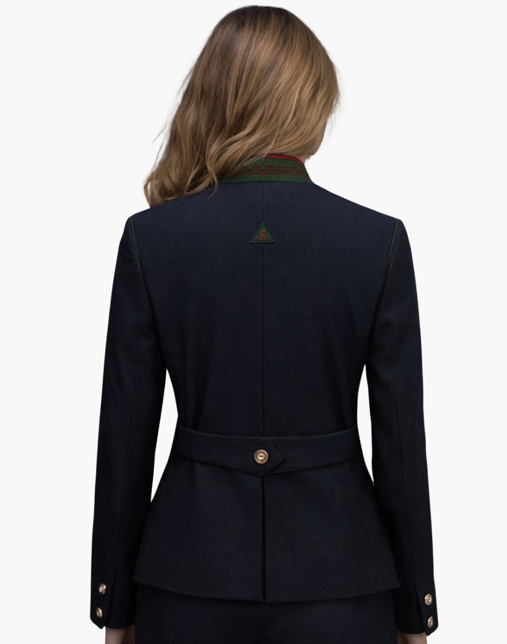 Back view of a woman wearing a fitted deep navy tailored jacket with a structured silhouette and a peplum-style hem. The jacket features a decorative green-trimmed collar, a small triangular detail at the upper back, and an adjustable back belt with a single button at the waist. She pairs it with dark trousers. The background is plain light gray in a studio setting.