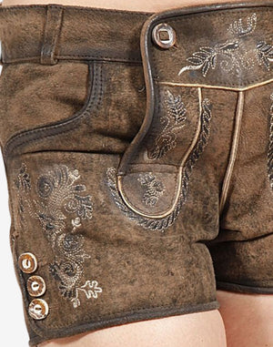 Close-up of rustic brown women’s suede lederhosen showing traditional embroidered floral patterns, decorative front flap detail, and side buttons on a distressed leather finish.