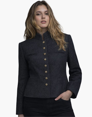 Front view of a woman wearing a dark charcoal tailored jacket with a stand-up collar and a vertical row of decorative gold-tone buttons down the front. The jacket features subtle welt pockets and structured seams that shape the waist, paired with dark jeans. The background is plain light gray in a clean studio setting.