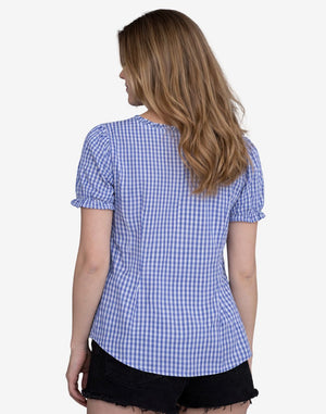 Back view of a woman wearing a blue and white gingham check dirndl blouse with short puff sleeves and a relaxed fit, paired with black denim shorts.