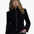 Front view of a woman wearing a black tailored jacket with a stand-up collar and a vertical row of decorative gold-tone buttons down the front. The jacket features subtle welt pockets and a gently flared hem, paired with dark jeans. The background is plain light gray, creating a clean studio look.