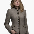 Front view of a woman wearing a tailored stone beige wool jacket with a stand-up collar and dark brown trim along the front opening and collar. A vertical row of dark buttons runs down the center, and angled welt pockets accent the waist. She is styled with dark jeans. The background is plain light gray, creating a clean studio look.