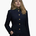 Front view of a woman wearing a deep navy tailored jacket with a stand-up collar accented in green and red trim. The jacket has a vertical row of gold-tone buttons, green-trimmed welt pockets, and a fitted silhouette, styled with dark trousers. The background is plain light gray.