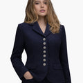 Front view of a woman wearing a deep navy tailored jacket with a notched collar and a vertical row of decorative silver-tone buttons down the front. The fitted jacket features flap pockets at the waist and structured seams for a shaped silhouette, paired with dark trousers. The background is plain light gray.