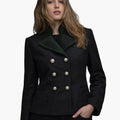 Front view of a woman wearing a dark wool tailored jacket with a wide green lapel collar featuring decorative dark appliqué details. The jacket has a double-breasted design with two vertical rows of large metallic buttons and subtle welt pockets, paired with dark jeans. The background is plain light gray, creating a clean studio look.