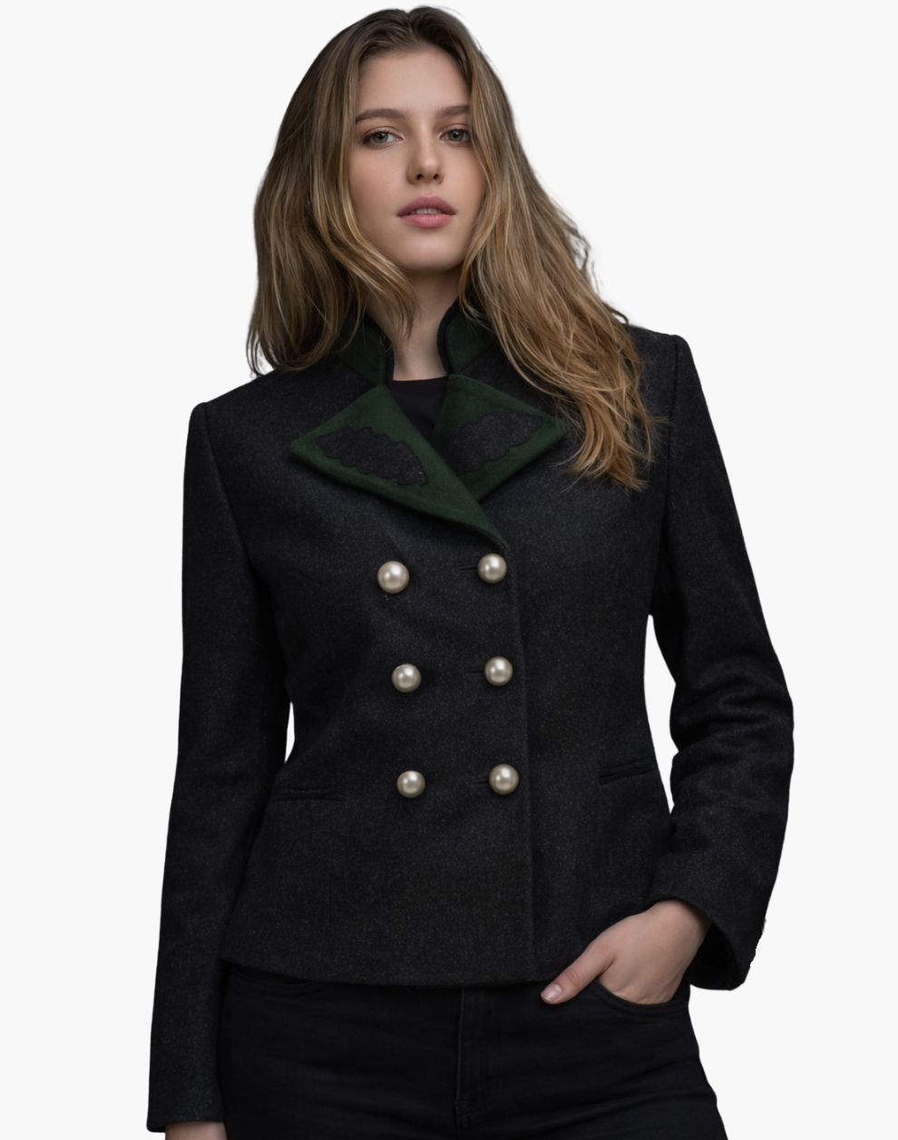 Front view of a woman wearing a dark wool tailored jacket with a wide green lapel collar featuring decorative dark appliqué details. The jacket has a double-breasted design with two vertical rows of large metallic buttons and subtle welt pockets, paired with dark jeans. The background is plain light gray, creating a clean studio look.