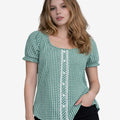 Front view of a woman wearing a green and white gingham dirndl-style blouse with short puff sleeves, decorative white lace panel and buttons, paired with black denim shorts.