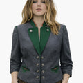 Woman wearing a grey Oktoberfest Trachten jacket with green trim, silver decorative buttons, and traditional Bavarian styling, paired with brown lederhosen, standing against a plain white background.