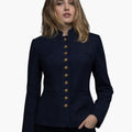 Front view of a woman wearing a deep navy tailored jacket with a stand-up collar and a vertical row of gold-tone buttons down the front. The fitted silhouette features subtle welt pockets and structured seams, styled with dark jeans. The background is plain light gray.