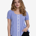Front view of a woman wearing a blue and white gingham dirndl blouse with short puff sleeves, lace trim and button detail, paired with black denim shorts.